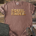 Fossil Finder Comfort Colors T-shirt in Espresso, laid flat. The words Fossil Finder are printed in cracked tan stone letters with ammonite shell fossils, plus a small ammonite after the last letter. Sleeves are rolled, on a rustic wood floor with olive cargo pants and brown hiking boots.