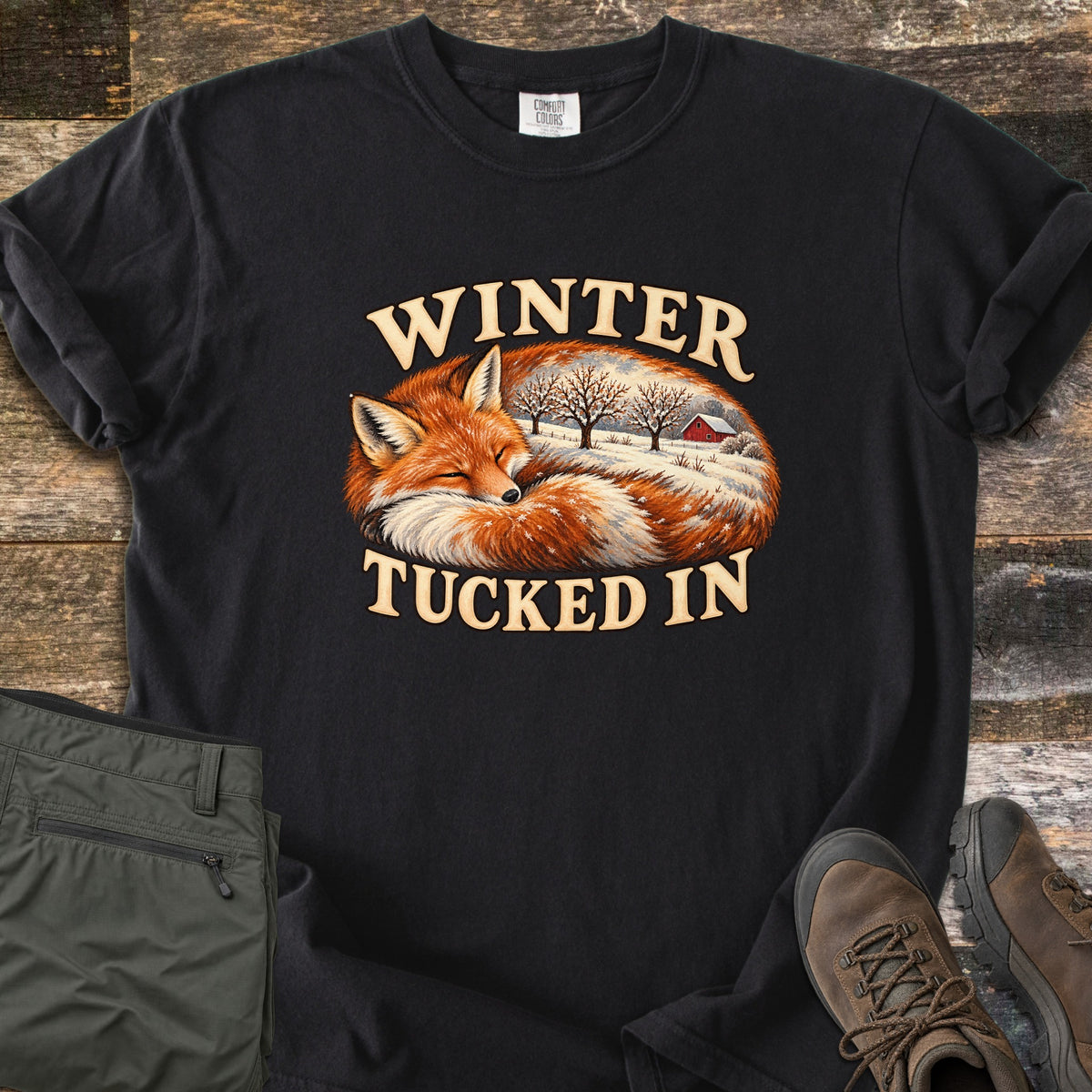 Winter Tucked In Comfort Colors T-shirt in Black, laid flat. A sleeping red fox curled up, with a snowy farm scene of bare trees and a red barn inside its body, between the words Winter Tucked In. Olive pants and brown hiking boots sit beside it on weathered wood.