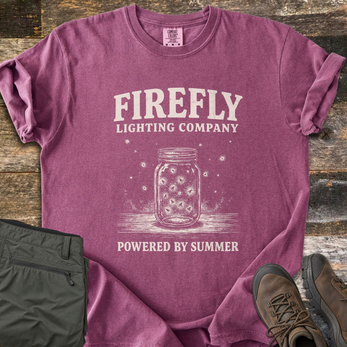 Firefly Lighting Company Comfort Colors T-shirt in Berry, laid flat. Cream print of a mason jar full of glowing fireflies on a wooden surface, under the words Firefly Lighting Company and above Powered by Summer. Olive cargo pants and brown hiking boots lie beside it on weathered wood.