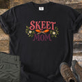 Skeet Mom Comfort Colors T-shirt in Black, laid flat. The words Skeet Mom in pink and plum lettering, with two orange clay targets crossed between them and sprigs of green leaves and white flowers on each side. Shown on weathered wood with olive pants and brown hiking boots.