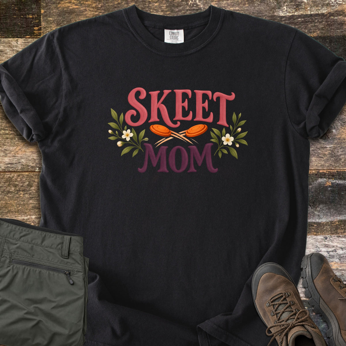 Skeet Mom Comfort Colors T-shirt in Black, laid flat. The words Skeet Mom in pink and plum lettering, with two orange clay targets crossed between them and sprigs of green leaves and white flowers on each side. Shown on weathered wood with olive pants and brown hiking boots.