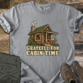 Grateful For Cabin Time Comfort Colors T-shirt in Grey, laid flat. Print of a log cabin with a green roof, smoking chimney, a pie on the porch rail and a pumpkin on the steps, above the words Grateful For Cabin Time. Green pants and brown hiking boots lie on weathered wood.