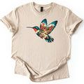 Hummingbird Canyon Comfort Colors T-shirt in Sand, laid flat. A hummingbird in flight with teal wings and a red throat, its body and wings filled with a desert canyon scene of red rock cliffs, a creek, shrubs and orange blooms. Sleeves cuffed, on a white surface.