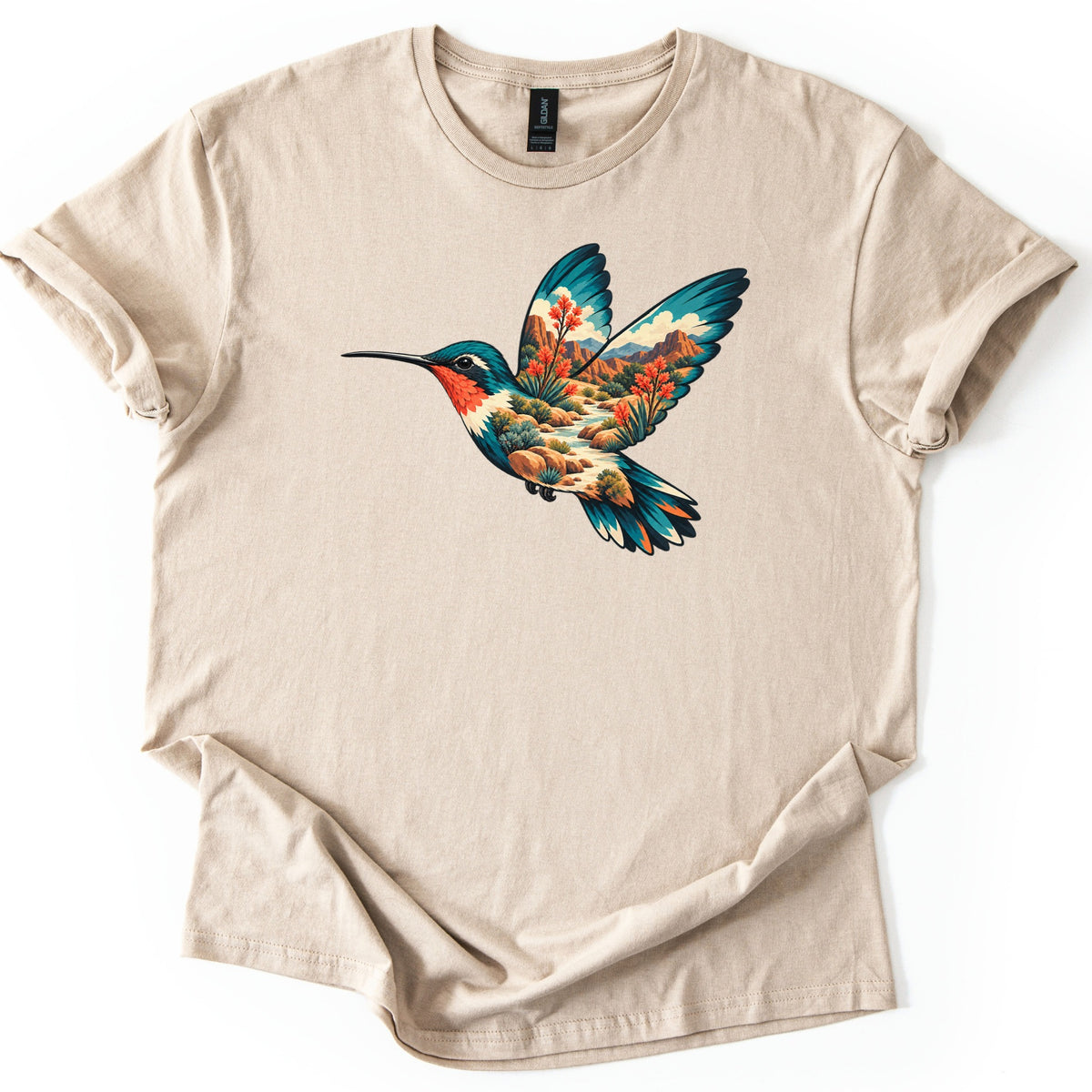 Hummingbird Canyon Comfort Colors T-shirt in Sand, laid flat. A hummingbird in flight with teal wings and a red throat, its body and wings filled with a desert canyon scene of red rock cliffs, a creek, shrubs and orange blooms. Sleeves cuffed, on a white surface.