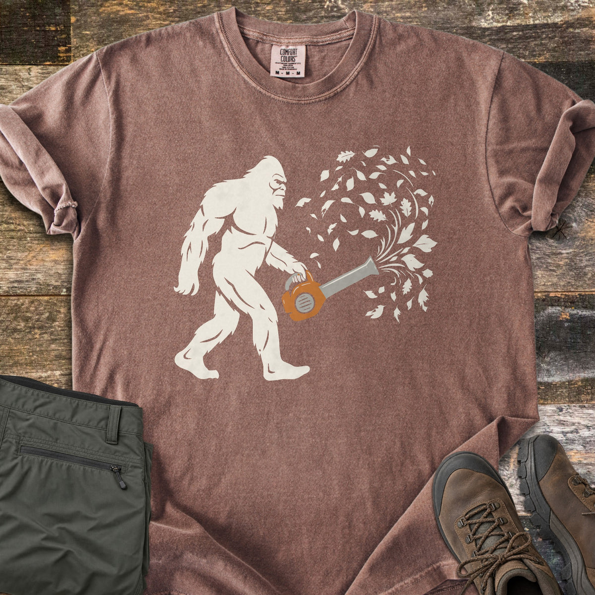 Bigfoot Leaf Blower Comfort Colors T-shirt in Espresso, laid flat. A white Bigfoot silhouette walks while holding an orange leaf blower that sends a swirl of white leaves into the air. Folded olive pants and brown hiking boots sit beside it on weathered wood boards.