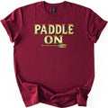 Paddle On Comfort Colors T-shirt in Maroon, laid flat. The words PADDLE ON in cream, weathered wood-style letters with orange riveted edges and green shadows sit above a wooden canoe paddle with green stripes. Cuffed sleeves, on a white background.