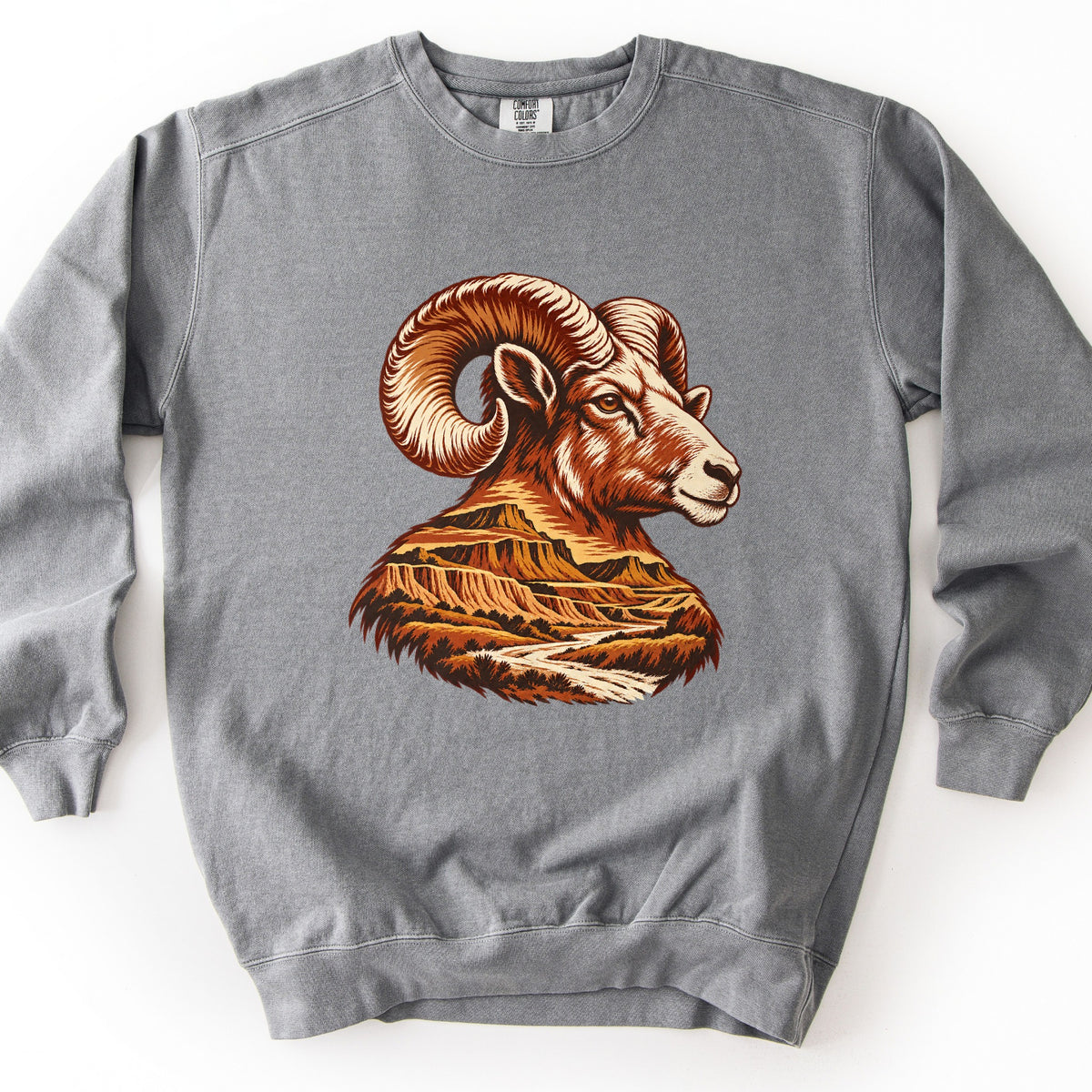 Bighorn Badlands Comfort Colors crewneck sweatshirt in Grey, laid flat. A woodcut-style bighorn ram head with curled horns, in rust, brown and cream, faces right; its chest becomes a badlands scene of layered buttes and a winding river. No words. Set on a white surface.