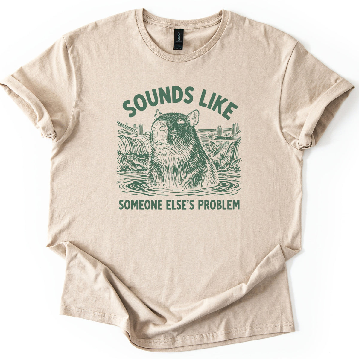 Sounds Like Someone Else's Problem Comfort Colors T-shirt in Sand, laid flat. A dark green sketch of a calm capybara with closed eyes sitting in a pond, with waterfalls and broken fences behind it, between the words Sounds Like and Someone Else's Problem. Cuffed sleeves, white background.
