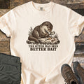 The Otter Has Seen Better Bait Comfort Colors T-shirt in Ivory, laid flat. A detailed brown illustration of an otter on a rock studying a fishing lure and a worm through a magnifying glass, above the words The Otter Has Seen Better Bait. Olive hiking pants and brown boots lie on wood planks.