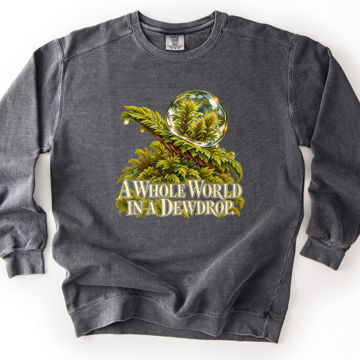 A Whole World in a Dewdrop Comfort Colors crewneck sweatshirt in Pepper, laid flat. A painted dewdrop rests on a green fern-like moss branch, the drop reflecting tiny evergreen sprigs, above cream lettering reading A Whole World in a Dewdrop. The sweatshirt lies on a white background.