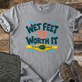 Wet Feet Worth It Comfort Colors T-shirt in Grey, laid flat. The words Wet Feet Worth It in dark teal with two orange footprints and blue water splashes above a yellow kayak and paddle. Laid on weathered wood with folded olive pants and brown hiking boots.