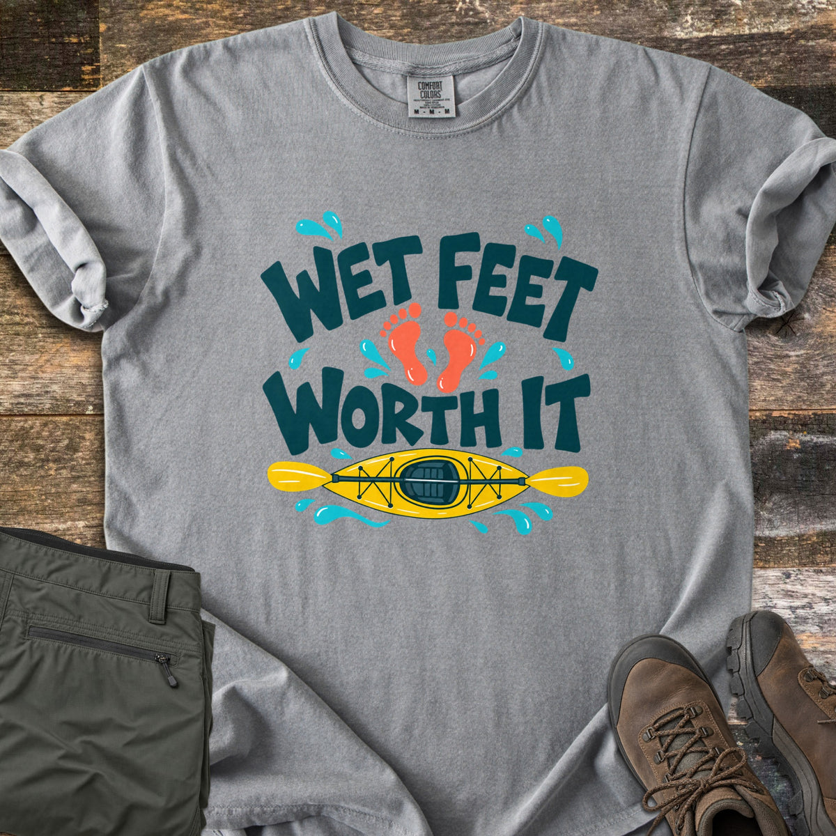 Wet Feet Worth It Comfort Colors T-shirt in Grey, laid flat. The words Wet Feet Worth It in dark teal with two orange footprints and blue water splashes above a yellow kayak and paddle. Laid on weathered wood with folded olive pants and brown hiking boots.