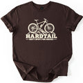 Hardtail Soft Spot Comfort Colors T-shirt in Dark Chocolate, laid flat. A cream-colored print of a hardtail mountain bike resting on rocks, above the words HARDTAIL and Soft Spot for Rocks. The shirt has rolled sleeves and lies on a white background.