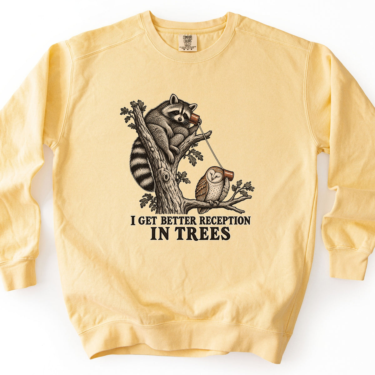 I Get Better Reception in Trees Comfort Colors crewneck sweatshirt in Butter, laid flat. A vintage-style etching of a raccoon in a tree holding a tin can phone to its ear, connected by string to a barn owl on a branch, above the words I Get Better Reception in Trees. Shown on a white background.