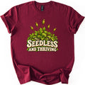 Seedless and Thriving Comfort Colors T-shirt in Maroon, laid flat. Green moss with tall reddish stalks topped by spore capsules grows over lichen-dotted rocks above the words SEEDLESS AND THRIVING in cream retro letters. The shirt lies on white with sleeves rolled.