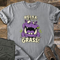 Relax He Eats Grass Comfort Colors T-shirt in Grey, laid flat. A grinning purple cartoon hippo with a sprig of green grass in its mouth sits between the words Relax, He Eats and Grass! Folded olive pants and brown hiking boots lie beside it on weathered wood planks.
