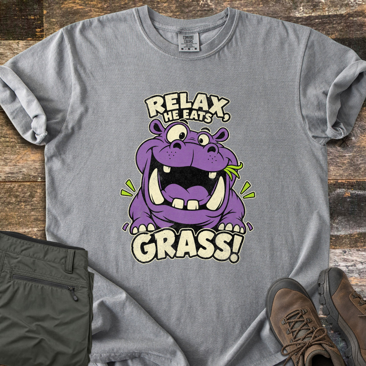 Relax He Eats Grass Comfort Colors T-shirt in Grey, laid flat. A grinning purple cartoon hippo with a sprig of green grass in its mouth sits between the words Relax, He Eats and Grass! Folded olive pants and brown hiking boots lie beside it on weathered wood planks.