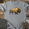 Prairie Bison Comfort Colors T-shirt in Grey, laid flat. A walking bison in black woodcut-style linework, its body filled with a golden prairie scene of grassland, a winding river and clouds. Folded olive cargo pants and brown hiking boots sit on weathered wood.