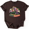 Mountain Bike Mom Raised on Switchbacks Comfort Colors T-shirt in Dark Chocolate, laid flat. Retro letters read Mountain Bike Mom under snowy peaks, an orange sun and pines, with a green and a smaller purple bike helmet on a winding dirt trail above Raised on Switchbacks. Sleeves rolled, on white.