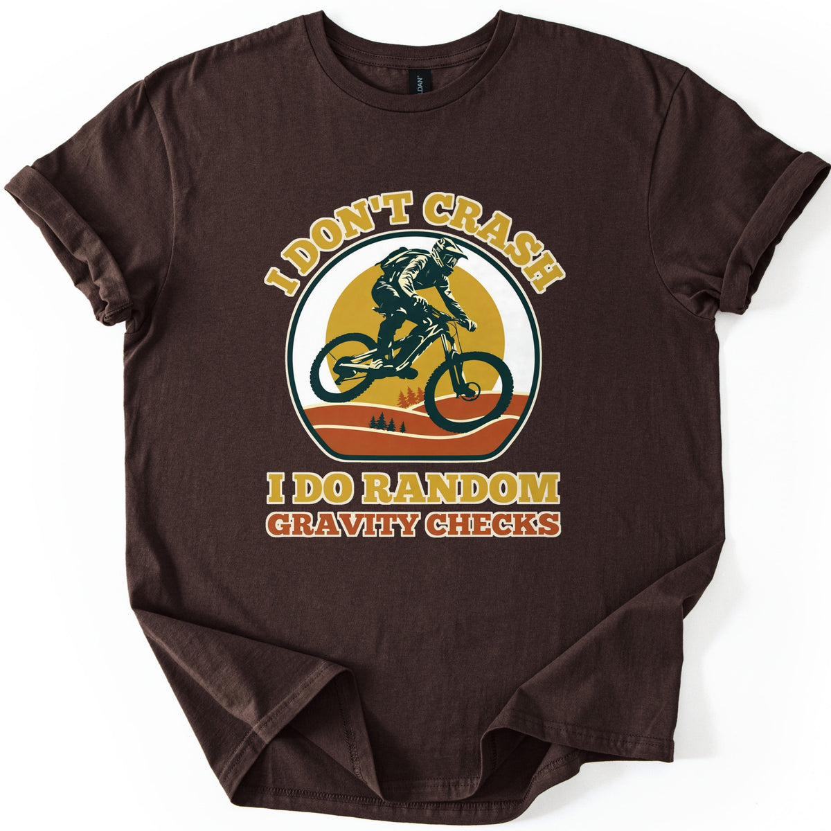 Random Gravity Checks MTB Comfort Colors T-shirt in Dark Chocolate, laid flat. A mountain biker in a full-face helmet jumps against a mustard sun over orange hills with pine trees, framed by the words I Don't Crash above and I Do Random Gravity Checks below. Sleeves rolled, on white.