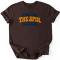 Here for the Spin Comfort Colors T-shirt in Dark Chocolate, laid flat. Retro wavy bubble letters read Here For in navy, The Spin in mustard yellow, and Helice in navy across the chest. The shirt has rolled sleeves and lies on a plain white background.