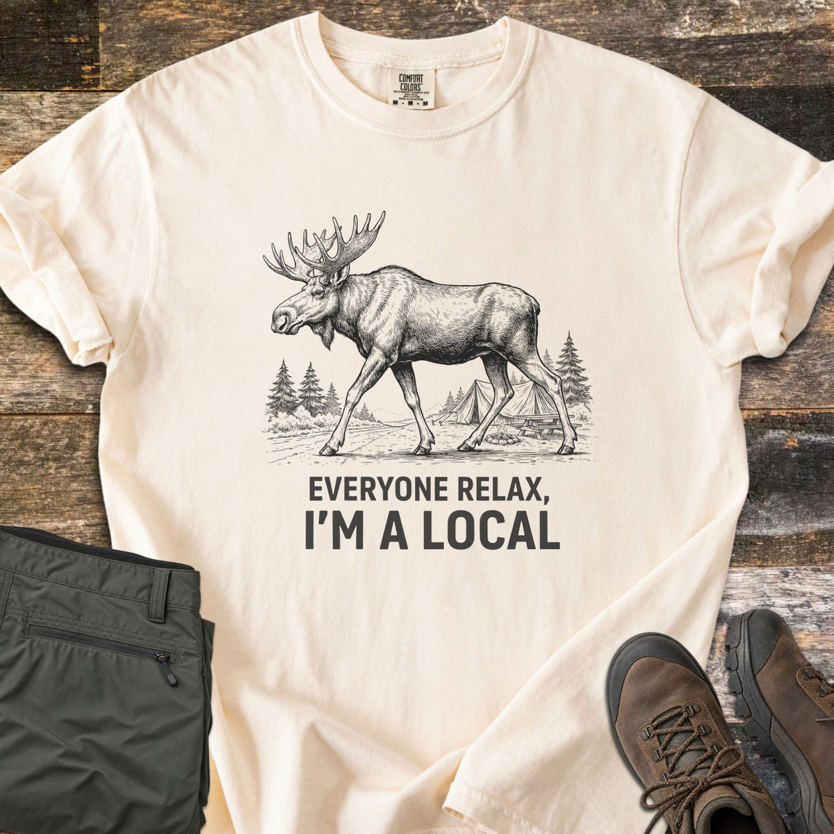 Everyone Relax I'm A Local Comfort Colors T-shirt in Ivory, laid flat. A sketched moose walks past pine trees and tents at a campsite above the words Everyone Relax, I'm A Local. Sleeves are rolled on a wood floor beside folded green pants and brown hiking boots.