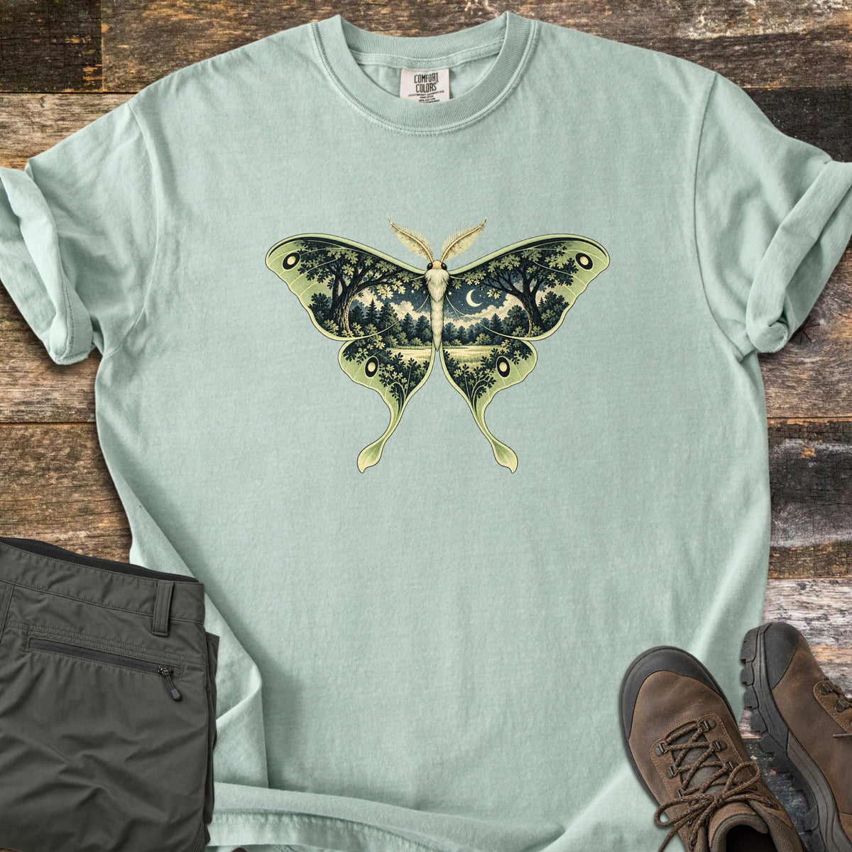Luna Moth Night Comfort Colors T-shirt in Bay, laid flat. A luna moth with pale green wings, each wing filled with a night forest scene of dark trees, a meadow and a crescent moon. Rolled sleeves; olive cargo pants and brown hiking boots lie beside it on weathered wood.