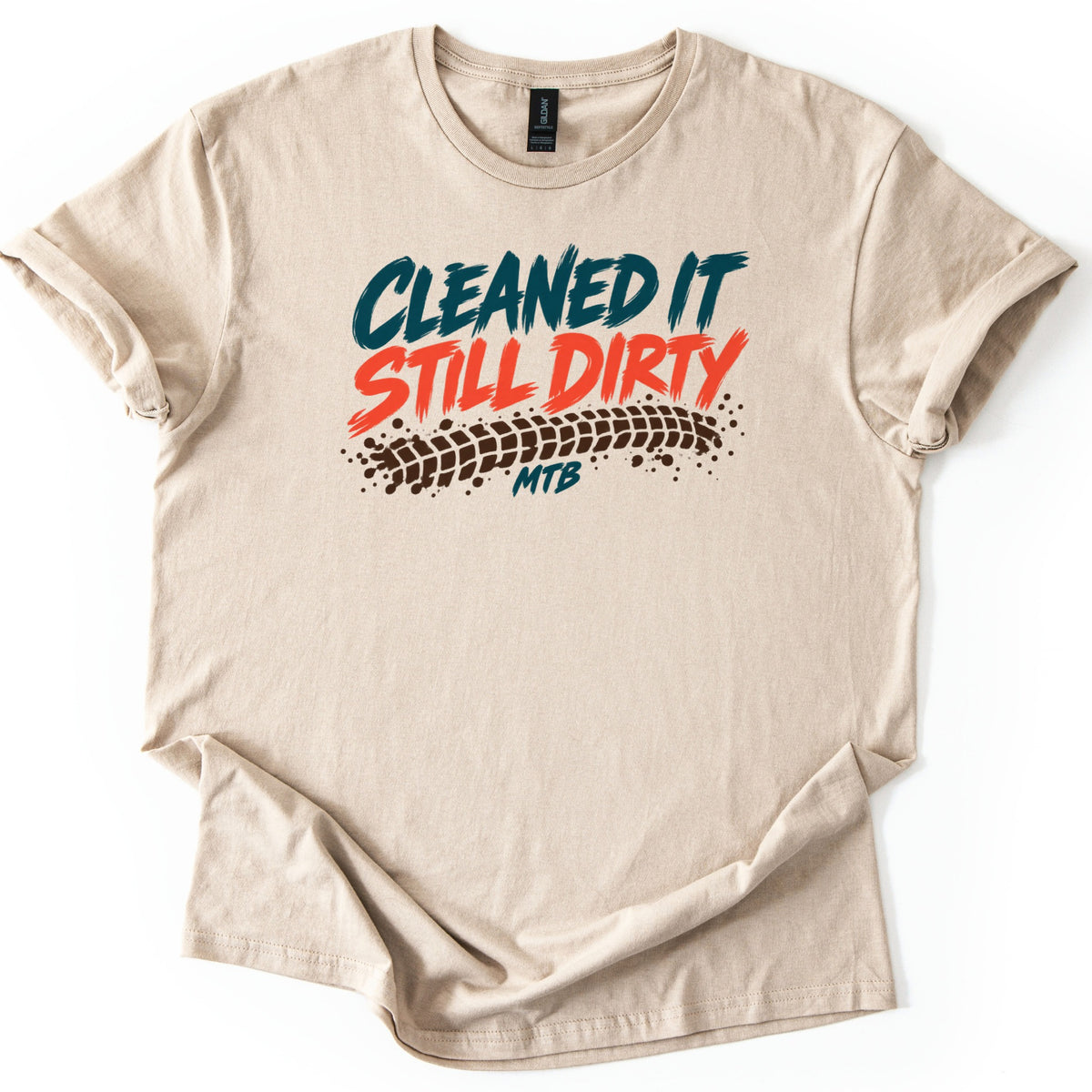 Cleaned It Still Dirty Comfort Colors T-shirt in Sand, laid flat. Brushstroke words Cleaned It in dark teal and Still Dirty in orange-red sit above a brown muddy tire track and the letters MTB. The shirt has rolled sleeves on a white background.