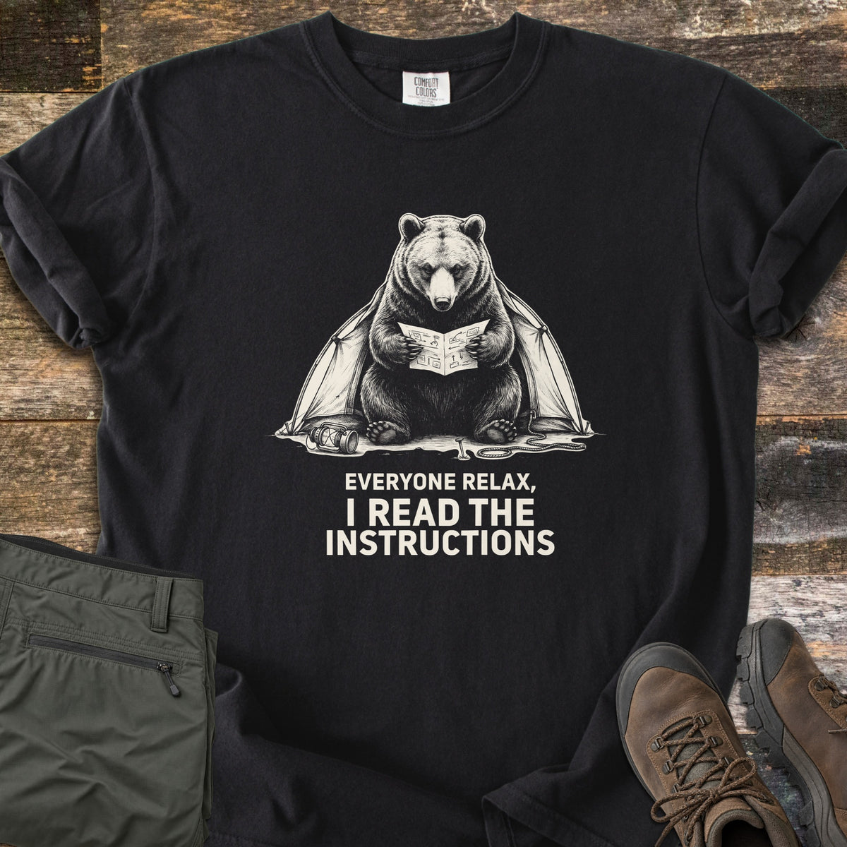 Everyone Relax I Read The Instructions Comfort Colors T-shirt in Black, laid flat. A cream sketch of a bear reading instructions in front of a tent, with a tipped lantern and rope, above the words Everyone relax, I read the instructions. Olive cargo pants and brown hiking boots sit on dark wood.