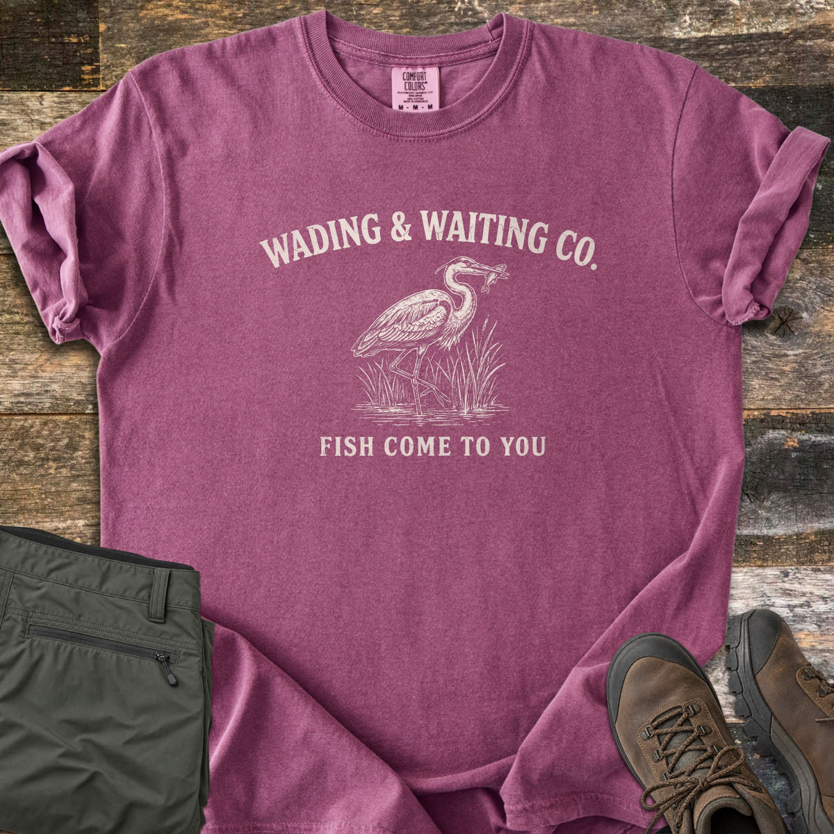 Wading & Waiting Co. Comfort Colors T-shirt in Berry, laid flat. A heron stands in marsh reeds holding a fish in its beak, under the arched words Wading & Waiting Co. and above Fish Come To You. Olive cargo pants and brown hiking boots lie beside it on weathered wood.