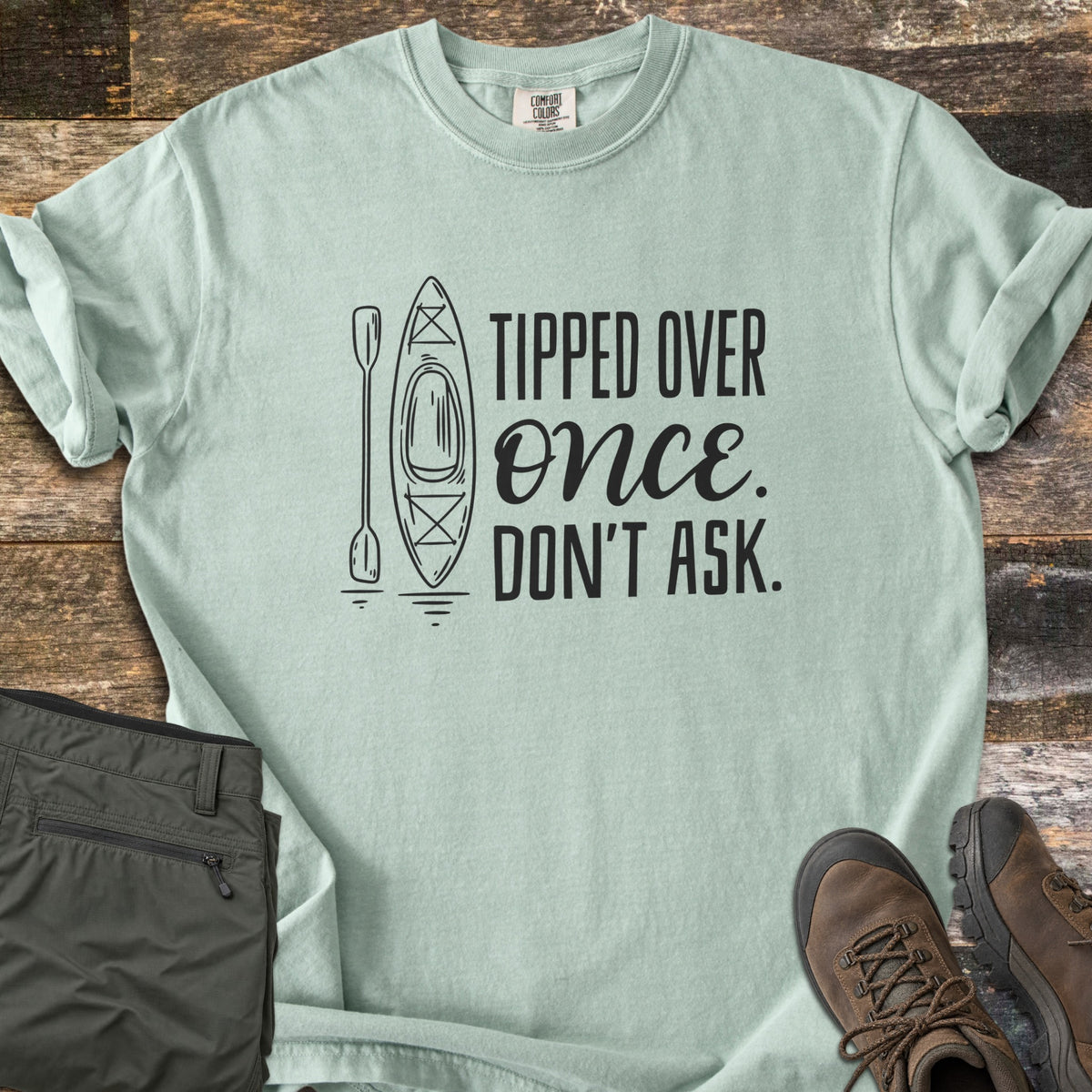 Tipped Over Once. Don't Ask. Comfort Colors T-shirt in Bay, laid flat. A black line drawing of a kayak and paddle sits beside the words Tipped Over Once. Don't Ask., with Once in script. Sleeves rolled, on weathered wood with folded olive hiking pants and brown hiking boots.