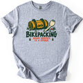 Bikepacking Room Service Is a Spork Comfort Colors T-shirt in Sport Grey, laid flat. A rolled yellow bedroll with green straps and a tent scene lies beside a spork above the words Bikepacking, Room Service Is a Spork, flanked by pine trees. The shirt has rolled sleeves on a white background.