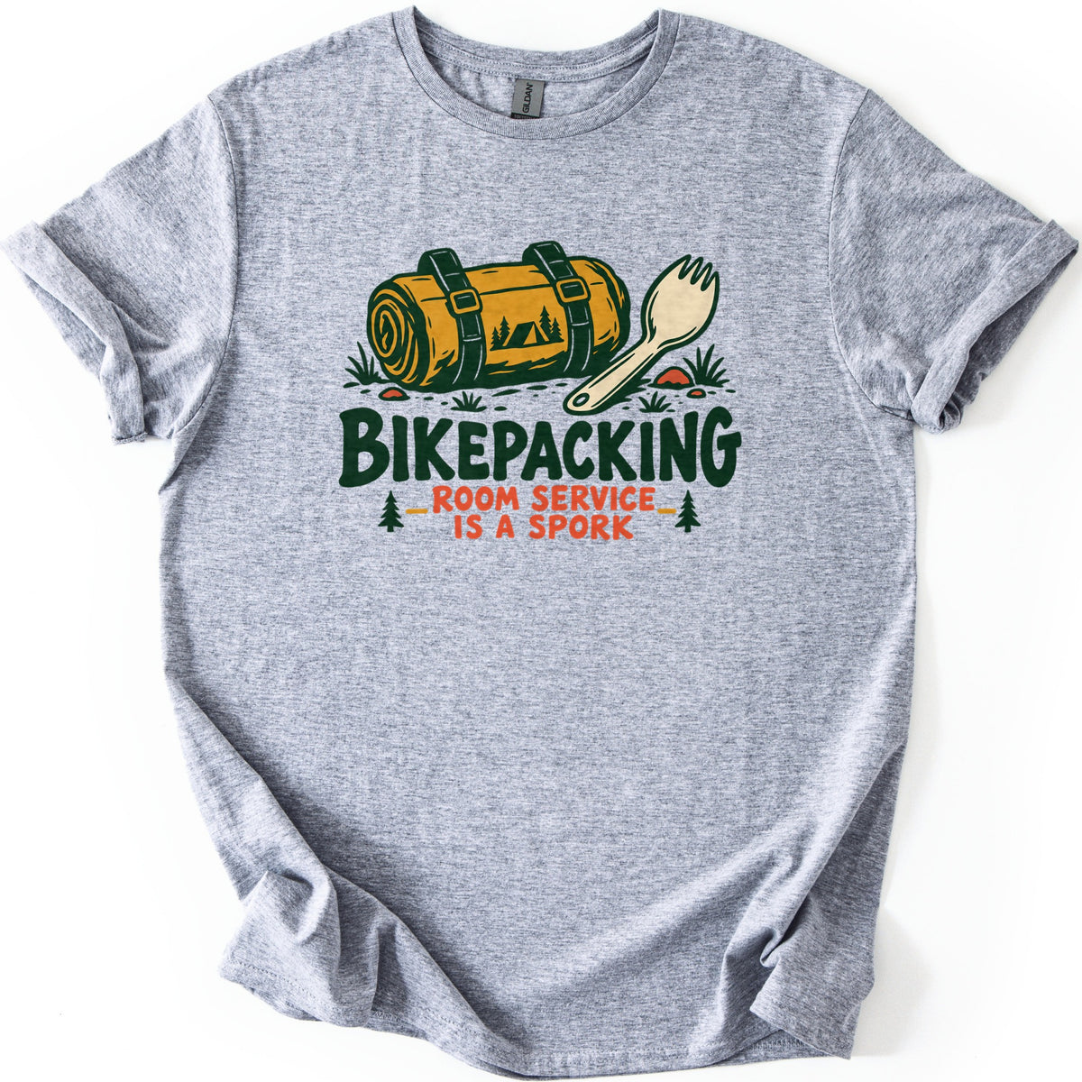 Bikepacking Room Service Is a Spork Comfort Colors T-shirt in Sport Grey, laid flat. A rolled yellow bedroll with green straps and a tent scene lies beside a spork above the words Bikepacking, Room Service Is a Spork, flanked by pine trees. The shirt has rolled sleeves on a white background.