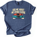 Other Kayak Comfort Colors T-shirt in Heather Navy, laid flat. Cream letters read Ask Me About My Other Kayak above a top view of an orange kayak, a longer teal kayak, and a cream double-bladed paddle. The shirt has rolled sleeves on a white background.