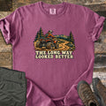 The Long Way Looked Better Comfort Colors T-shirt in Berry, laid flat. A rider in an orange jacket and helmet drives a red ATV down a winding dirt trail past pine trees and mountains, above the words The Long Way Looked Better. Olive pants and brown hiking boots sit on weathered wood.