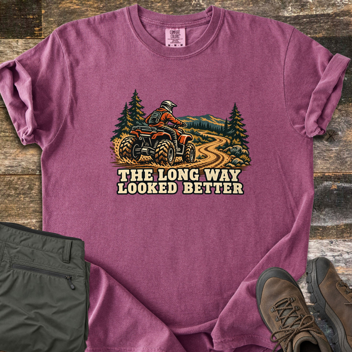 The Long Way Looked Better Comfort Colors T-shirt in Berry, laid flat. A rider in an orange jacket and helmet drives a red ATV down a winding dirt trail past pine trees and mountains, above the words The Long Way Looked Better. Olive pants and brown hiking boots sit on weathered wood.