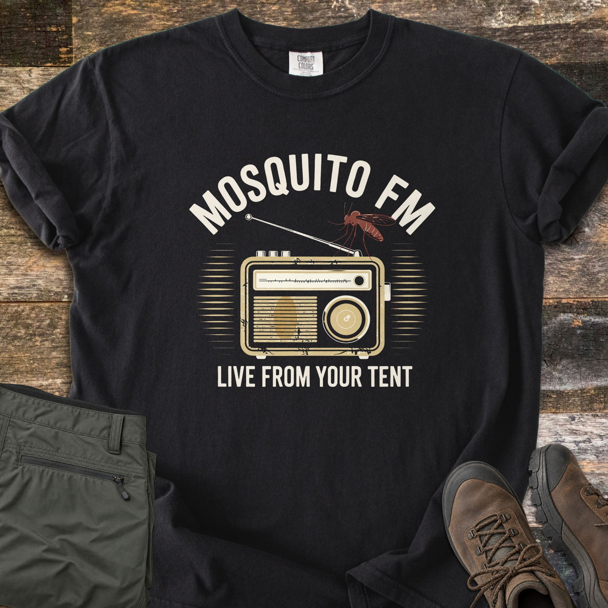 Mosquito FM Comfort Colors T-shirt in Black, laid flat. A mosquito perches on the antenna of a vintage tan radio under the arched cream words Mosquito FM, with Live From Your Tent below. Set on weathered wood with folded olive hiking pants and brown hiking boots.