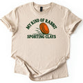 My Kind of Rabbit Comfort Colors T-shirt in Sand, laid flat. An orange clay target rolls through grass with motion lines, framed by the arched words My Kind of Rabbit above and Sporting Clays below in dark green. The shirt has rolled sleeves on a white background.