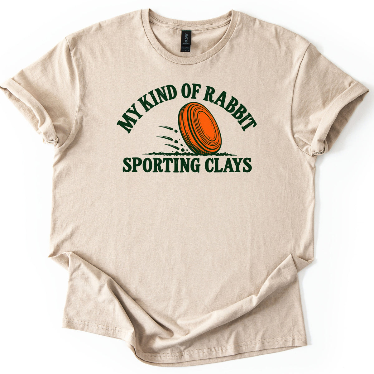 My Kind of Rabbit Comfort Colors T-shirt in Sand, laid flat. An orange clay target rolls through grass with motion lines, framed by the arched words My Kind of Rabbit above and Sporting Clays below in dark green. The shirt has rolled sleeves on a white background.