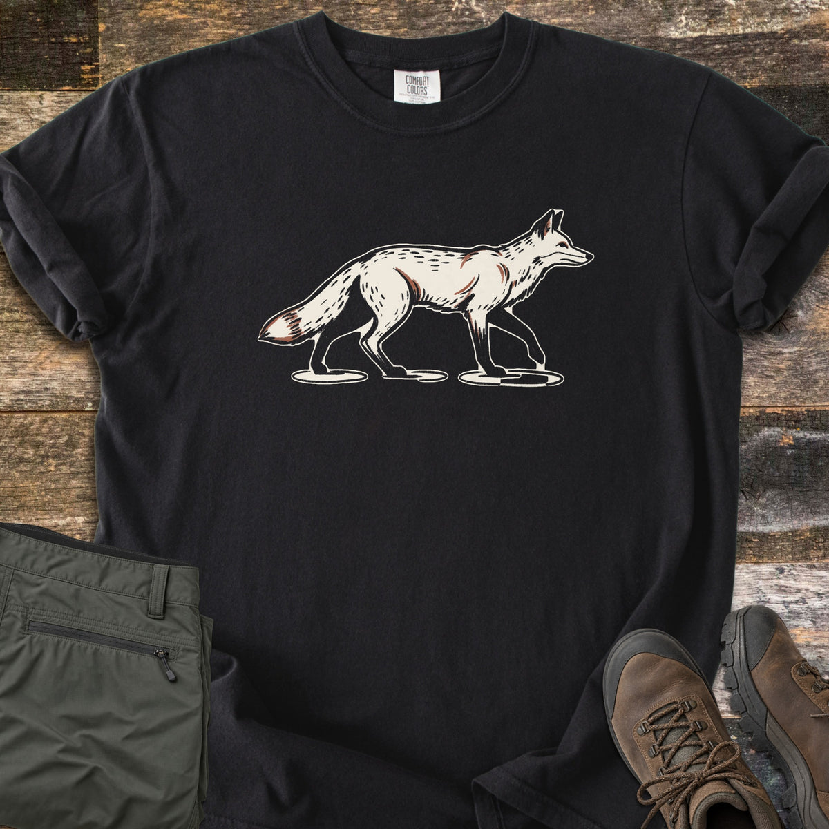 Trotting Fox Comfort Colors T-shirt in Black, laid flat. A cream and rust illustration of a fox trotting to the right across small ground patches, printed on the chest with no words. The shirt lies on weathered wood beside olive green hiking pants and brown hiking boots.