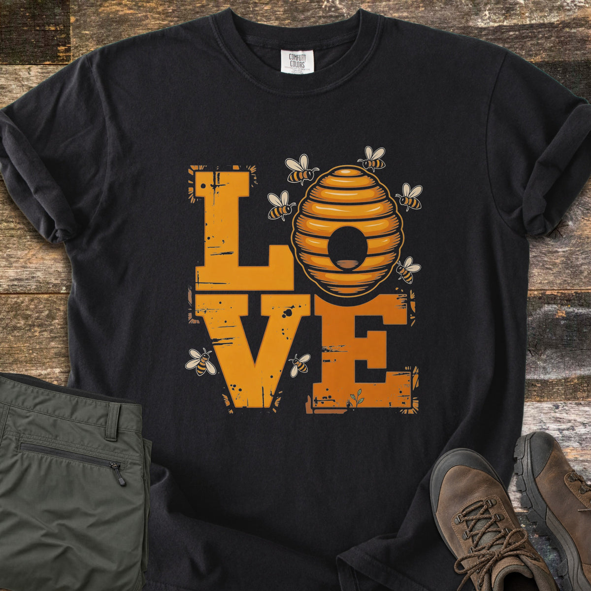 Beehive Love Comfort Colors T-shirt in Black, laid flat. The word LOVE in distressed orange block letters, with a striped beehive as the O and small bees flying around it. Olive cargo pants and brown hiking boots sit beside it on weathered wood boards.