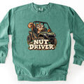 Nut Driver Comfort Colors crewneck sweatshirt in Light Green, laid flat. A grinning cartoon squirrel drives a muddy camo side-by-side off-road vehicle with an acorn on the hood, above the words Nut Driver in cream block letters. The sweatshirt lies on a white surface.