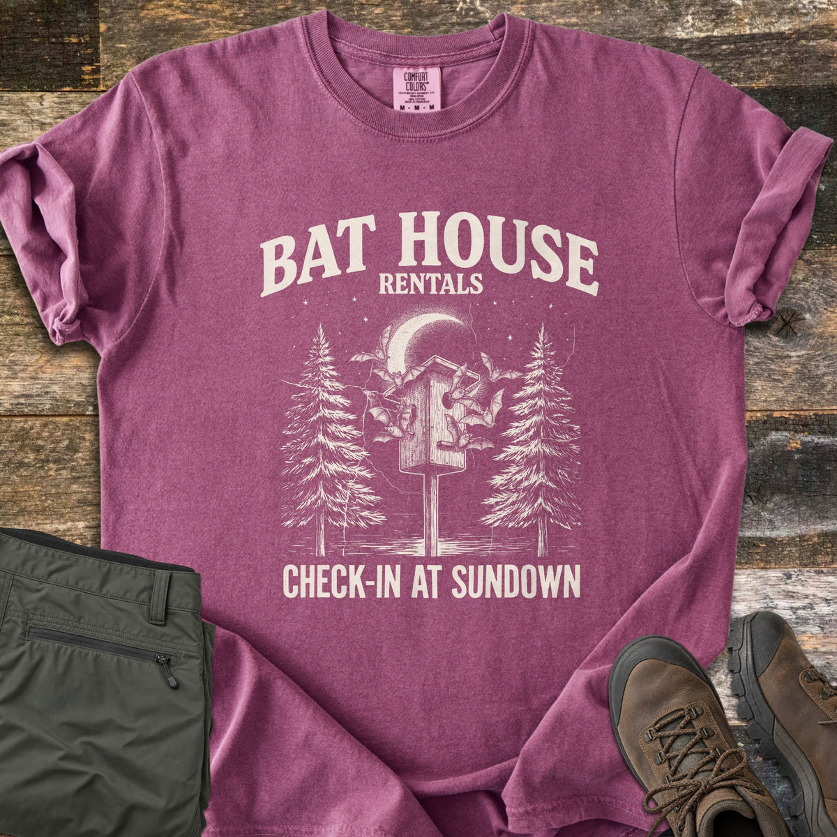 Bat House Rentals Comfort Colors T-shirt in Berry, laid flat. Off-white print of a bat house on a post with bats flying around it under a crescent moon, between two pine trees, with the words Bat House Rentals and Check-In at Sundown. Olive pants and brown hiking boots lie on wood planks.