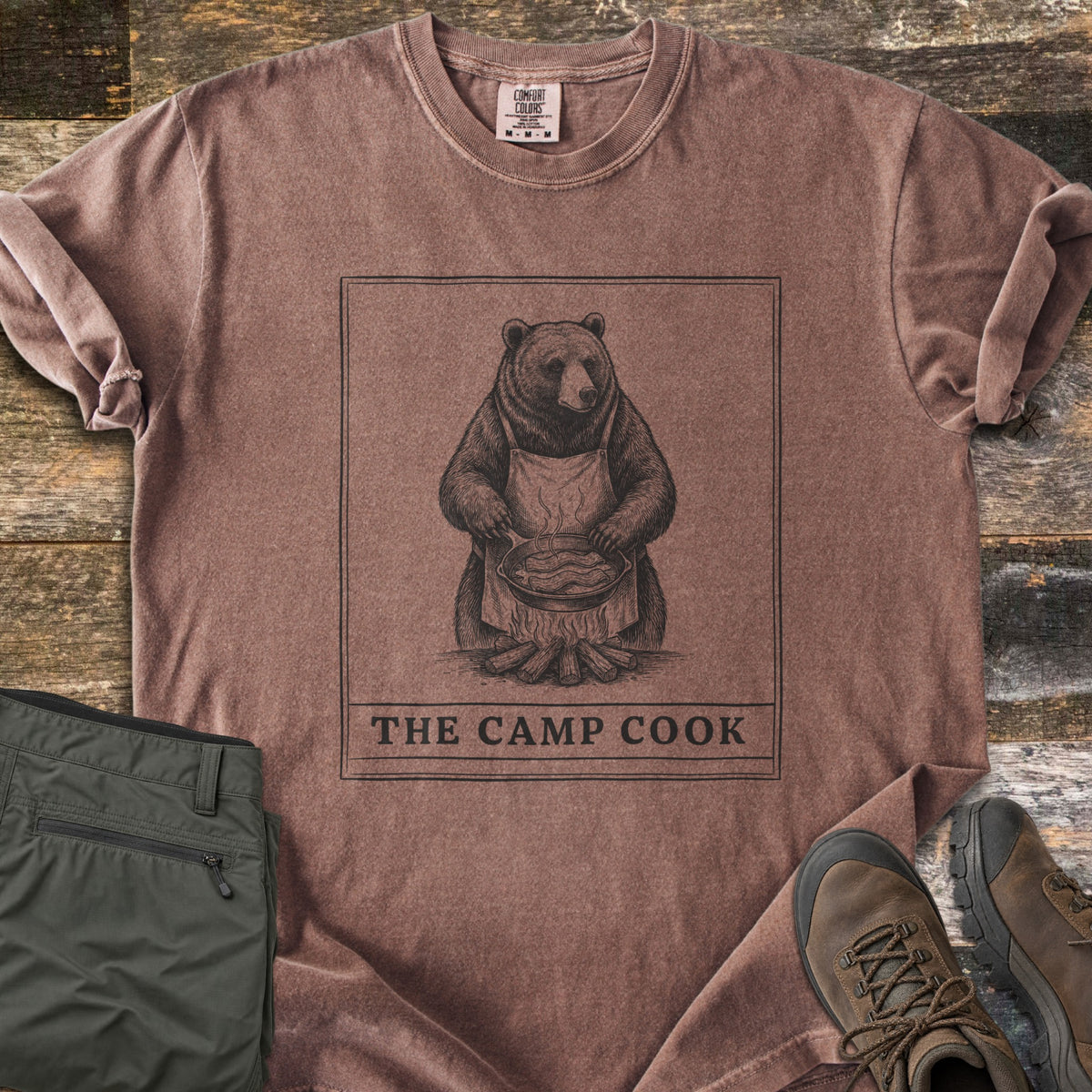 The Camp Cook Comfort Colors T-shirt in Espresso, laid flat. A framed sketch of a bear in an apron frying bacon in a skillet over a campfire, above the words THE CAMP COOK. Sleeves are cuffed on weathered wood beside olive hiking pants and brown hiking boots.