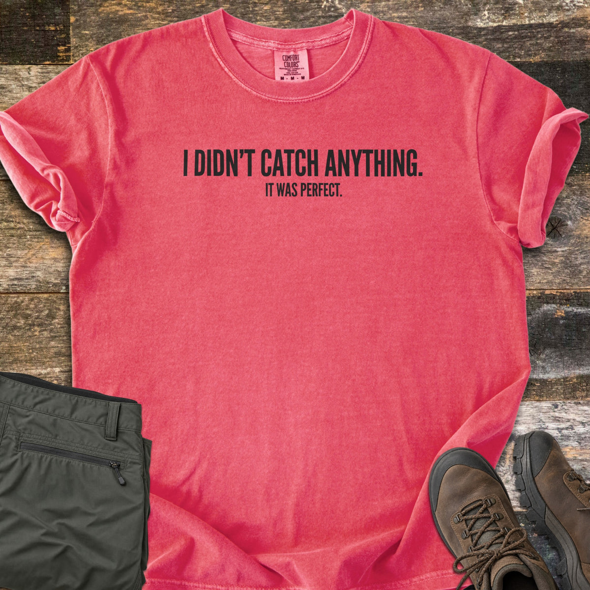 Didn't Catch Anything Comfort Colors T-shirt in Watermelon, laid flat. Black block letters read I Didn't Catch Anything. with It Was Perfect. in smaller letters below. Sleeves are rolled, with folded olive hiking pants and brown hiking boots on weathered wood planks.