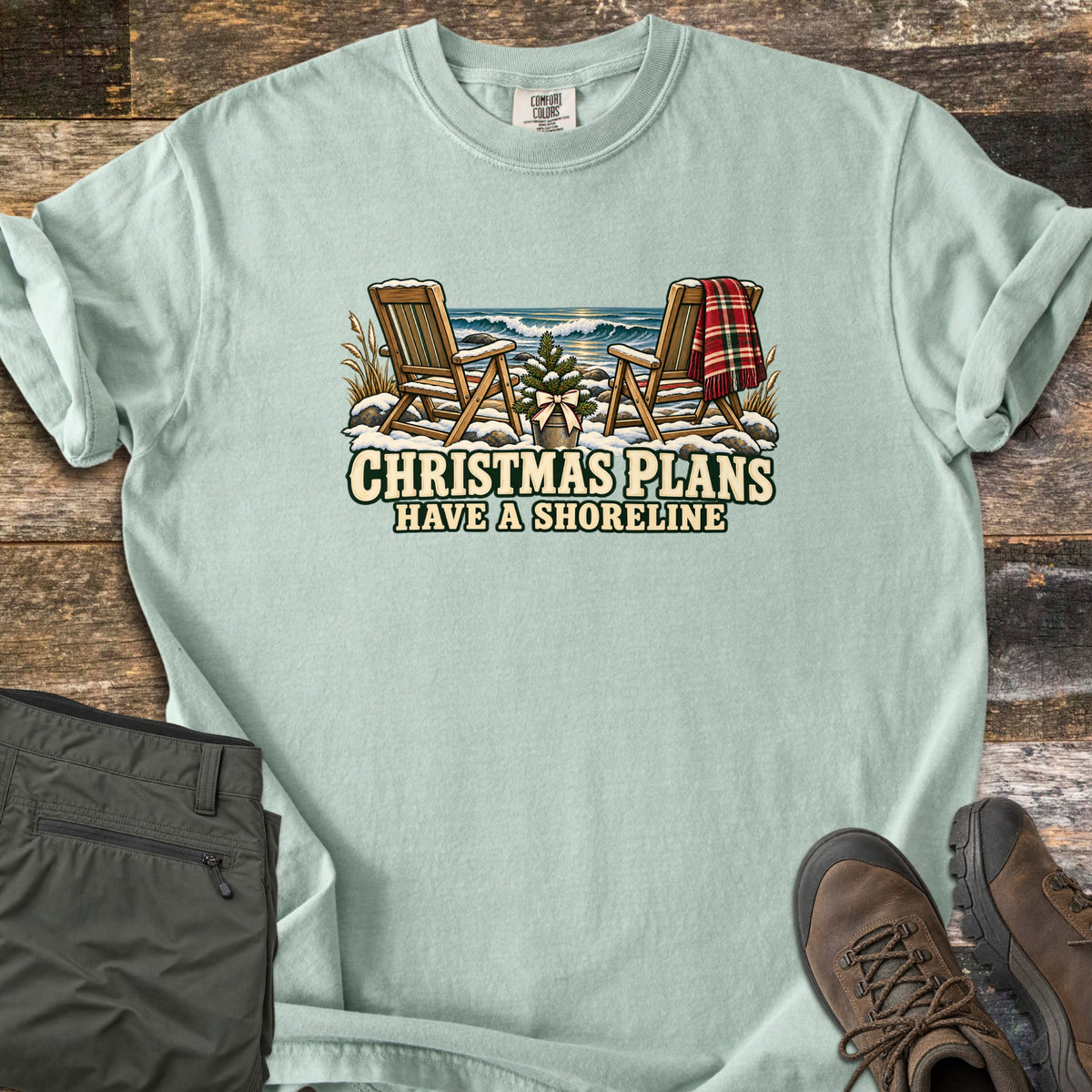 Christmas Plans Have a Shoreline Comfort Colors T-shirt in Bay, laid flat. Two snow-dusted wooden Adirondack chairs, one with a red plaid blanket, flank a small potted tree with a bow before ocean waves, above the words Christmas Plans Have a Shoreline. Olive pants and brown boots lie on weathered wood.