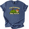 Two Kingdoms One Patch Comfort Colors T-shirt in Heather Navy, laid flat. A smiling green moss clump with two orange spore stalks sits beside a smiling spotted brown mushroom, between the words Two Kingdoms and One Patch. The sleeves are rolled on a white background.