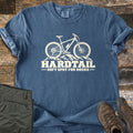 Hardtail Soft Spot Comfort Colors T-shirt in Denim, laid flat. A cream mountain bike with a water bottle on its frame sits on rocks above the words HARDTAIL SOFT SPOT FOR ROCKS. Sleeves rolled, on weathered wood planks beside folded olive pants and brown hiking boots.