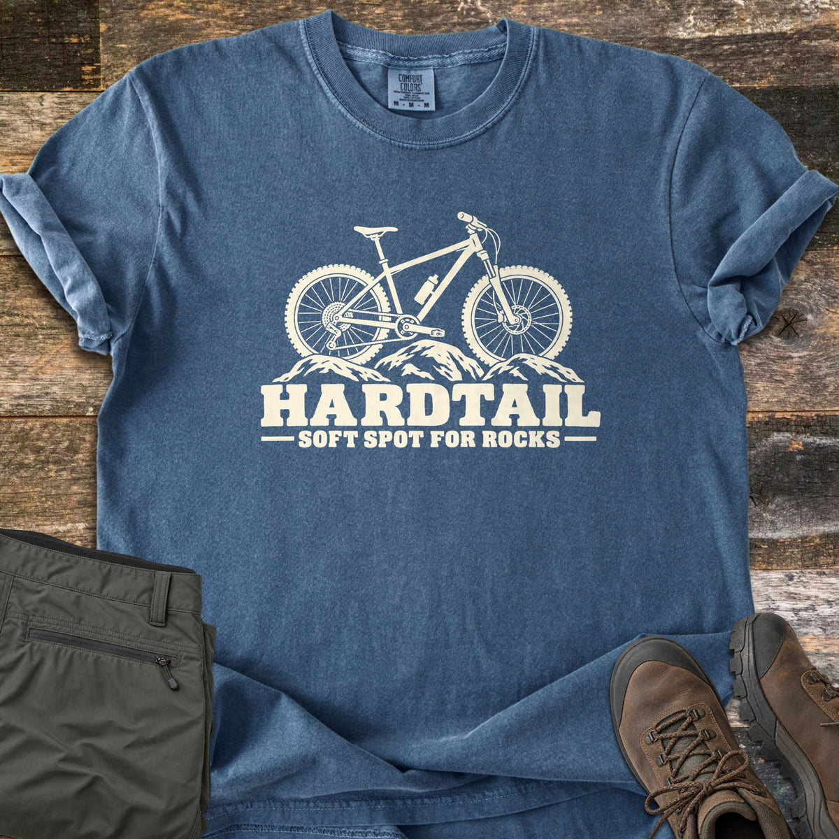 Hardtail Soft Spot Comfort Colors T-shirt in Denim, laid flat. A cream mountain bike with a water bottle on its frame sits on rocks above the words HARDTAIL SOFT SPOT FOR ROCKS. Sleeves rolled, on weathered wood planks beside folded olive pants and brown hiking boots.
