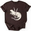 Otter In The Reeds Comfort Colors T-shirt in Dark Chocolate, laid flat. A cream silhouette of an otter with open mouth, whiskers and raised paws curls behind two tall reeds above swirling water lines. No words. The shirt lies on a white surface with sleeves rolled.
